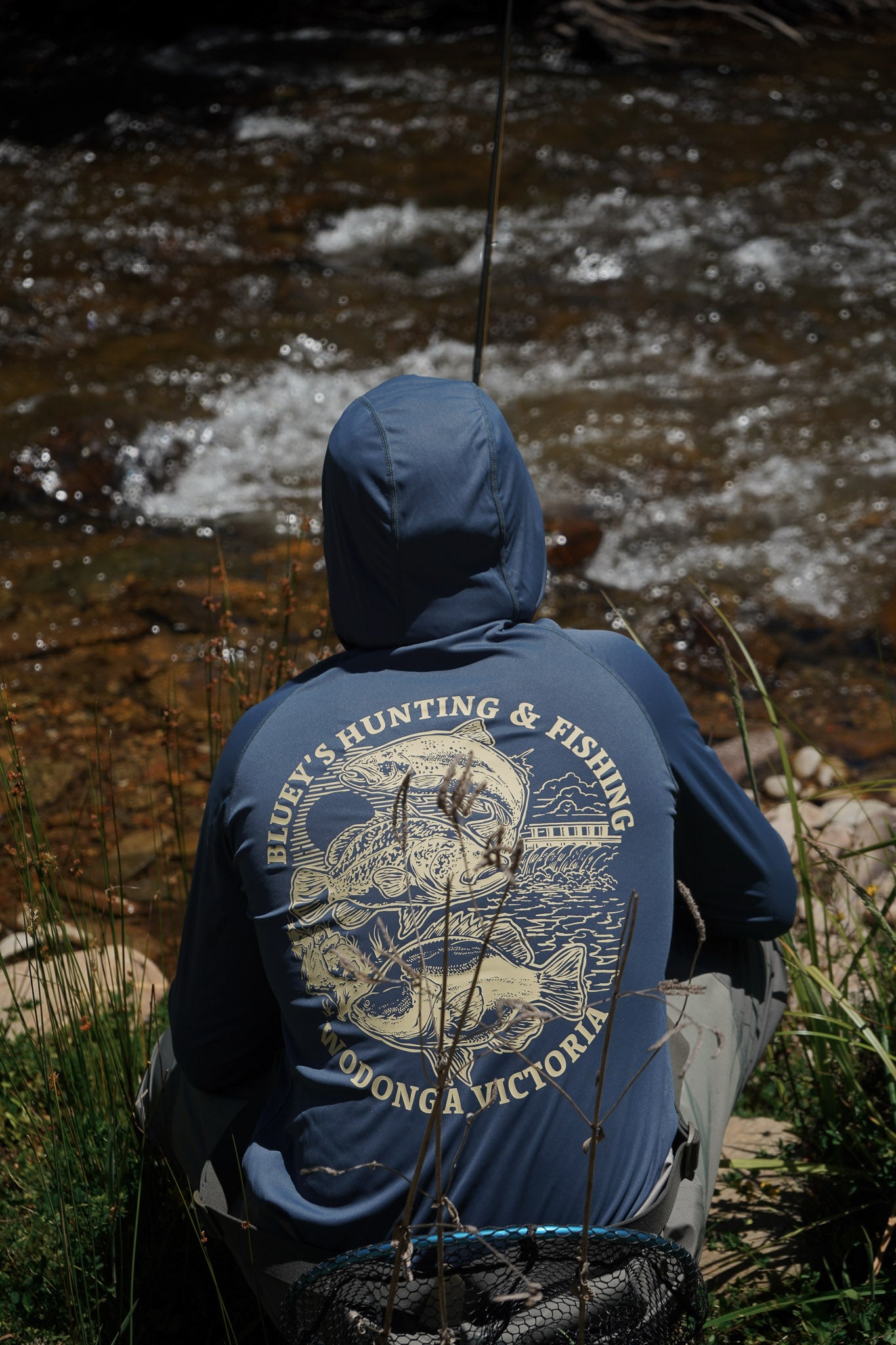 Person wearing a blue hoodie with hunting and fishing design by a river